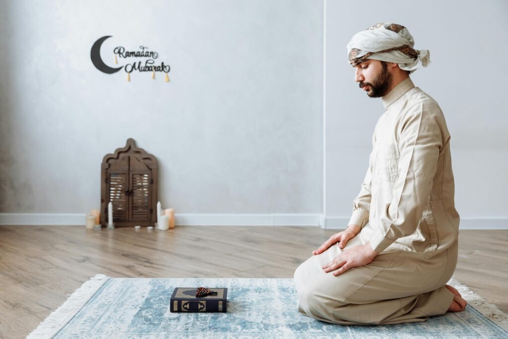 A man kneels in prayer on a rug during Ramadan in traditional clothing, with a Quran and decor.